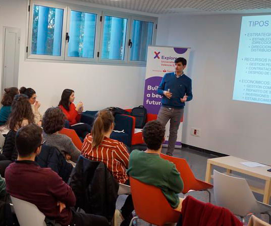 Hombre de pie delante de una pantalla impartiendo una charla a un grupo de personas sentadas delante